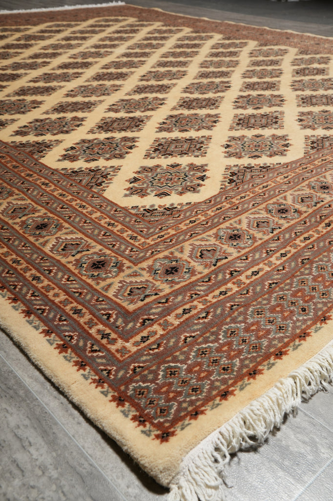 A close-up of a patterned area rug with cream, rust red, and brown tones, showing its detailed geometric design and fringed edge on a gray floor.
