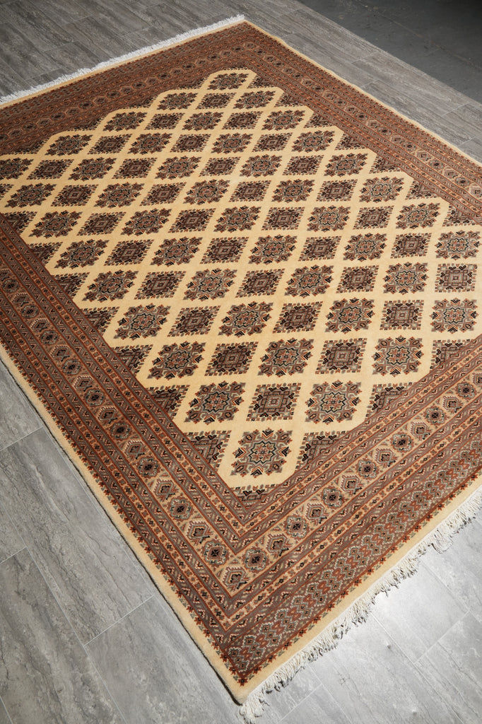 A rectangular area rug with a traditional Persian-style geometric pattern, featuring a large diamond lattice in the center and detailed borders. The rug has a warm brown and beige color palette with white fringe on the ends, shown laid out on a gray wood floor.