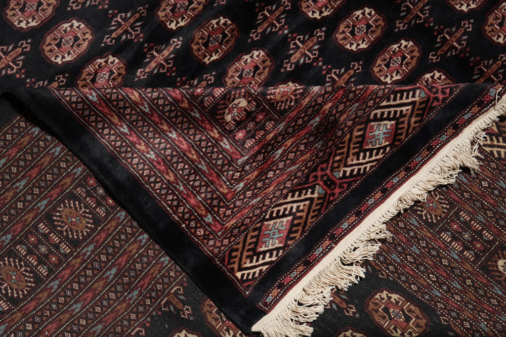 A close-up of a traditional patterned rug with a black base, intricate red, beige, and brown geometric motifs, and cream fringe on one corner.