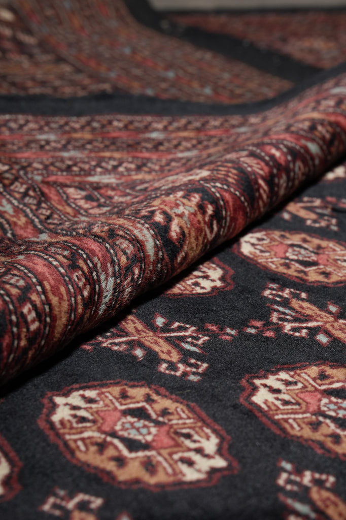 A close-up of a folded area rug showing its detailed pattern and pile texture. The rug features a dark charcoal black base with intricate traditional motifs in rust red, beige, and brown.