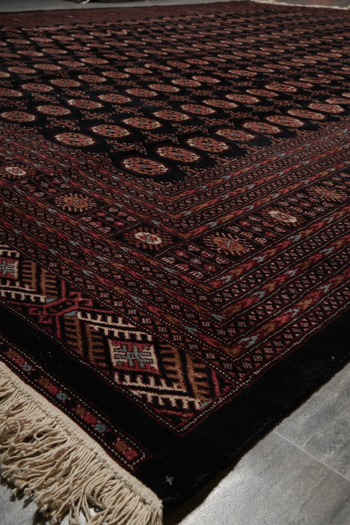 A close-up of a traditional patterned rug with a dark red and black base, featuring geometric borders, circular motifs, and cream-colored fringe on one end, laid on a gray tiled floor.
