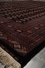 Load image into Gallery viewer, A close-up of a traditional patterned rug with a dark red and black base, featuring geometric borders, circular motifs, and cream-colored fringe on one end, laid on a gray tiled floor.