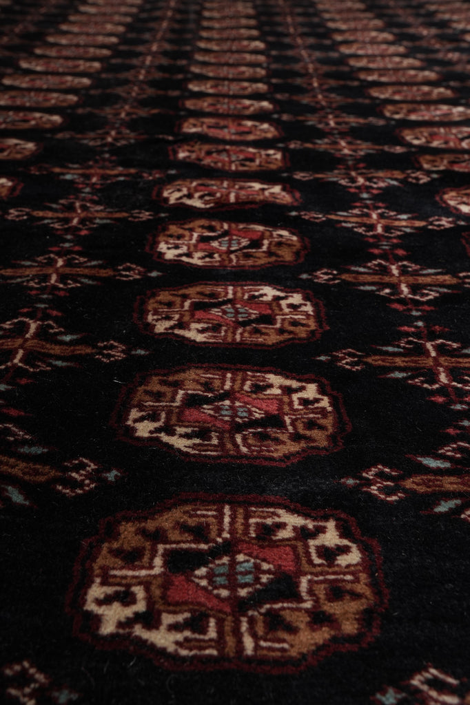 A close-up of a dark patterned rug or textile with repeating ornate medallion motifs in red, beige, and brown on a black background.