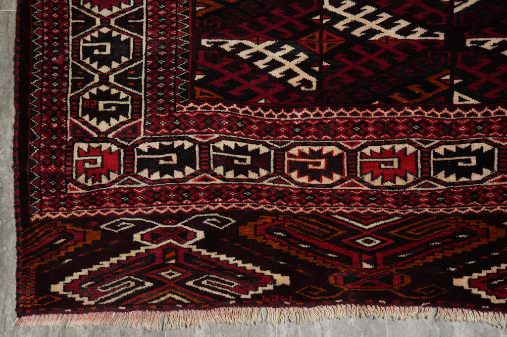 A close-up of a patterned rug corner showing geometric and tribal-inspired motifs in deep red, black, beige, and brown, with fringed edges visible at the bottom.