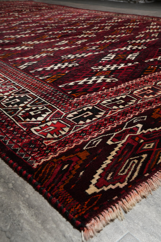 A close-up view of a traditional patterned rug with a deep red and black base, cream and beige geometric motifs, and fringed edges.