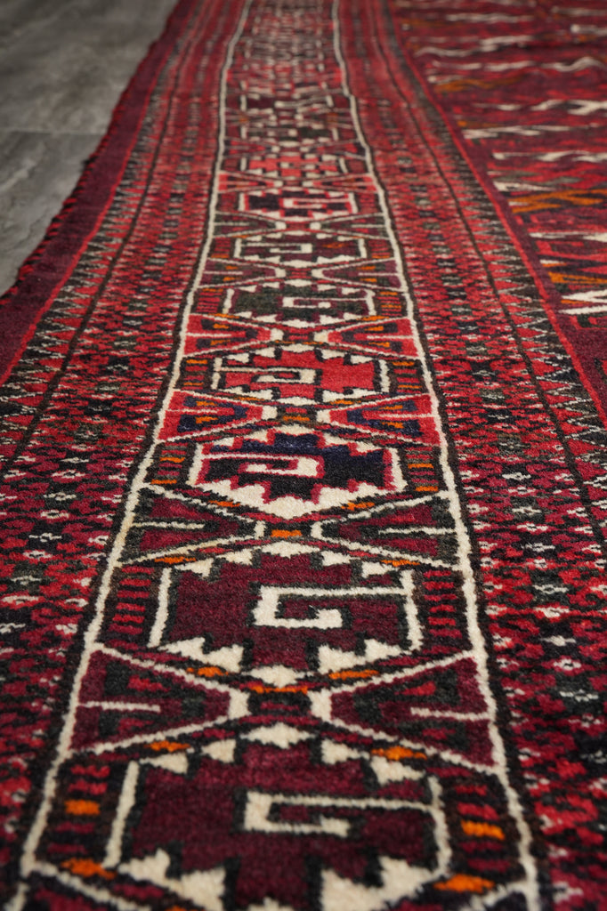 A close-up view of a traditional patterned area rug with a geometric design in red, black, cream, and orange, laid out on a gray floor.