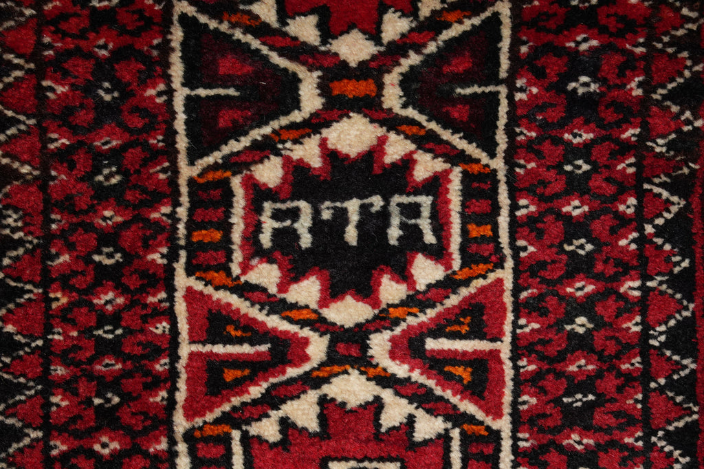 A close-up of a patterned rug featuring a central geometric motif with the letters 'RTA' in white on a black background, surrounded by red, black, and cream patterns.