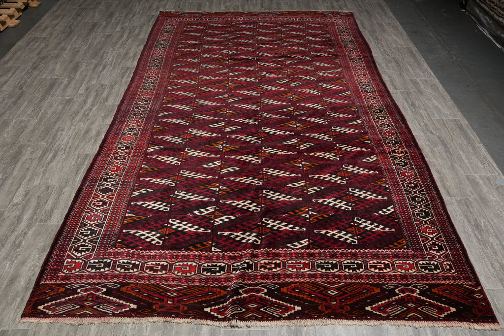 A traditional Persian-style wool rug with a dark red field, cream and black geometric patterns, and a detailed border, laid out on a gray wood floor.
