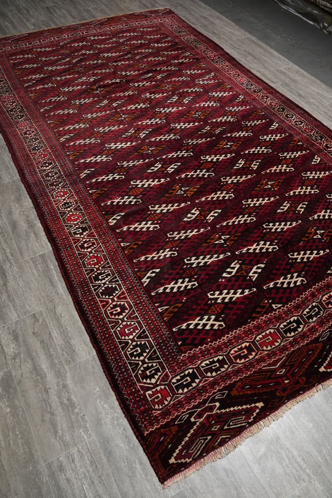 A traditional Persian-style area rug with a deep red field, featuring a repeating diamond pattern in cream, black, and red, and a detailed border.