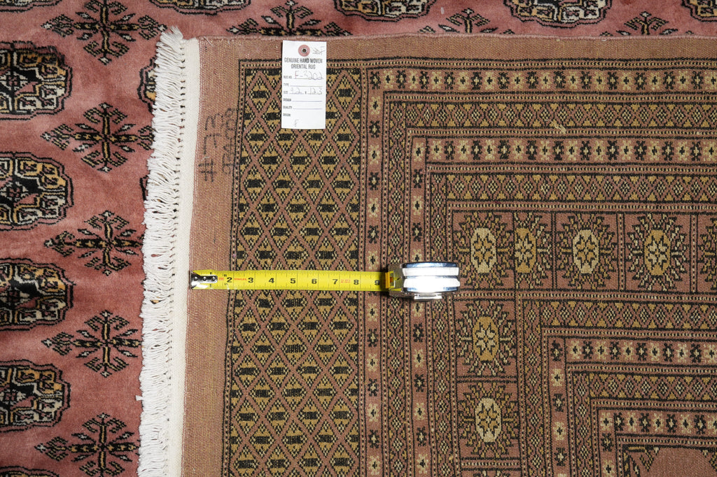 A patterned area rug with a dusty rose and brown geometric design, featuring white fringe on one side. A yellow tape measure is laid horizontally across the rug to show its dimensions, and a white product tag is visible near the top.