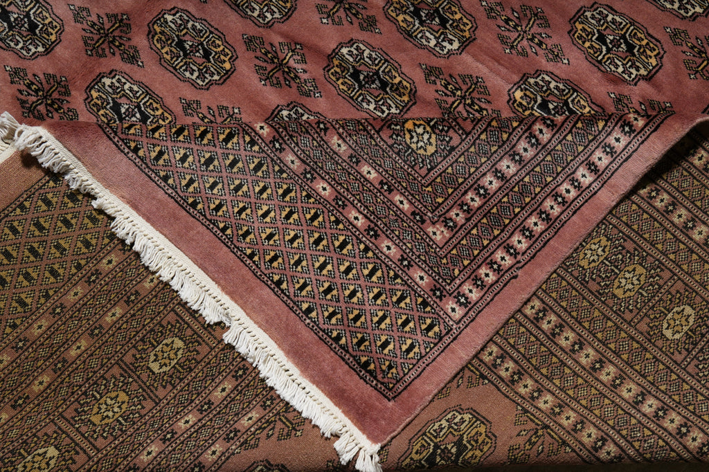 A close-up of a patterned area rug with a dusty rose pink field, intricate black, brown, and gold geometric motifs, and white fringe on one corner, laid over another matching rug.