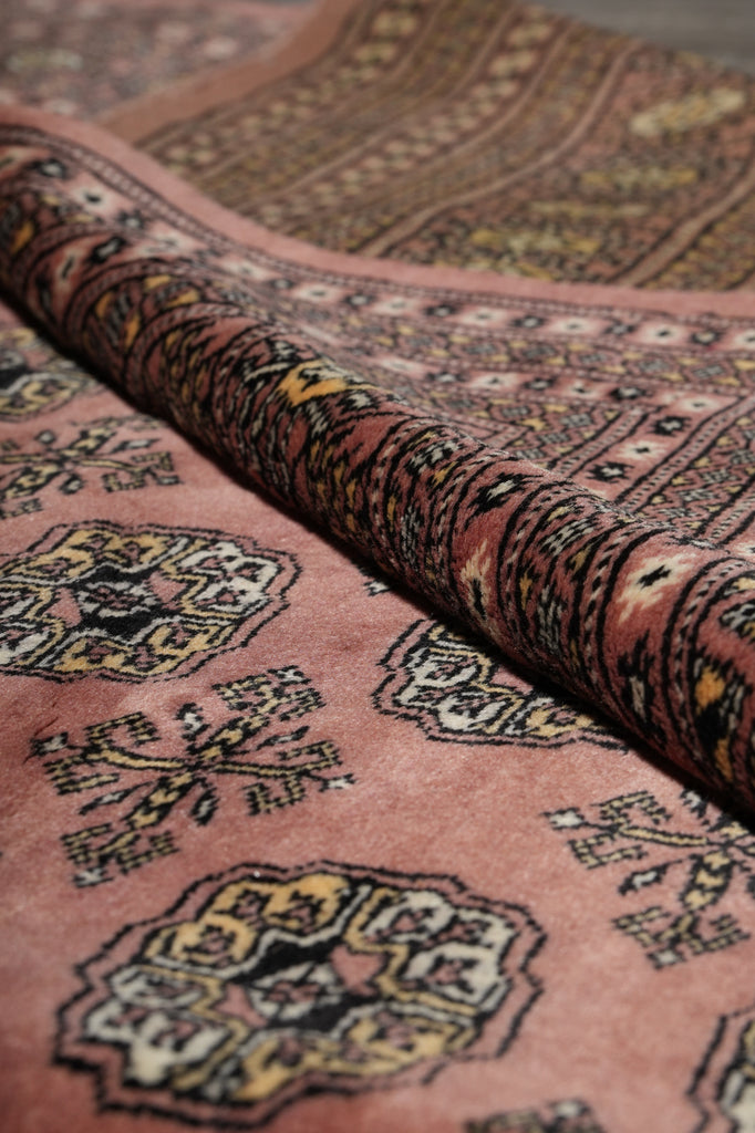 A close-up of a folded patterned rug showing a dusty pink field with ornate geometric and floral motifs in black, beige, and muted yellow, plus a dark patterned border.