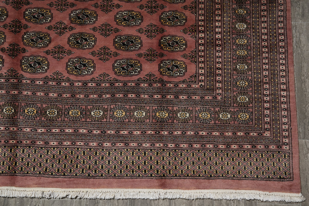 A close-up of an oriental rug showing its detailed pattern, featuring a dusty rose field with black, gold, and beige motifs, a dark patterned border, and white fringe at the bottom.