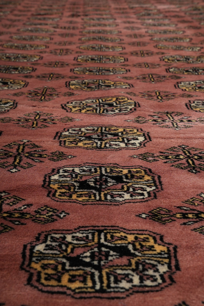 A close-up of a patterned rug with a dusty rose or terracotta pink field, featuring repeating geometric medallion motifs in black, yellow, and cream.
