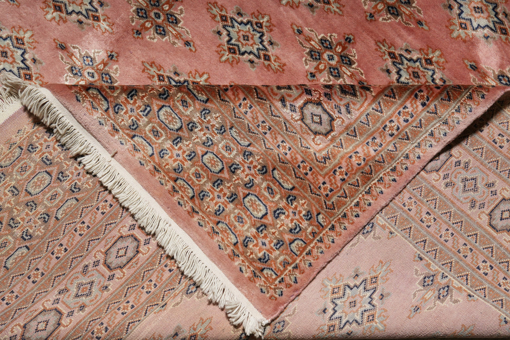A folded dusty pink patterned rug with cream fringe, featuring traditional geometric and floral motifs in muted blue, beige, and rust tones.