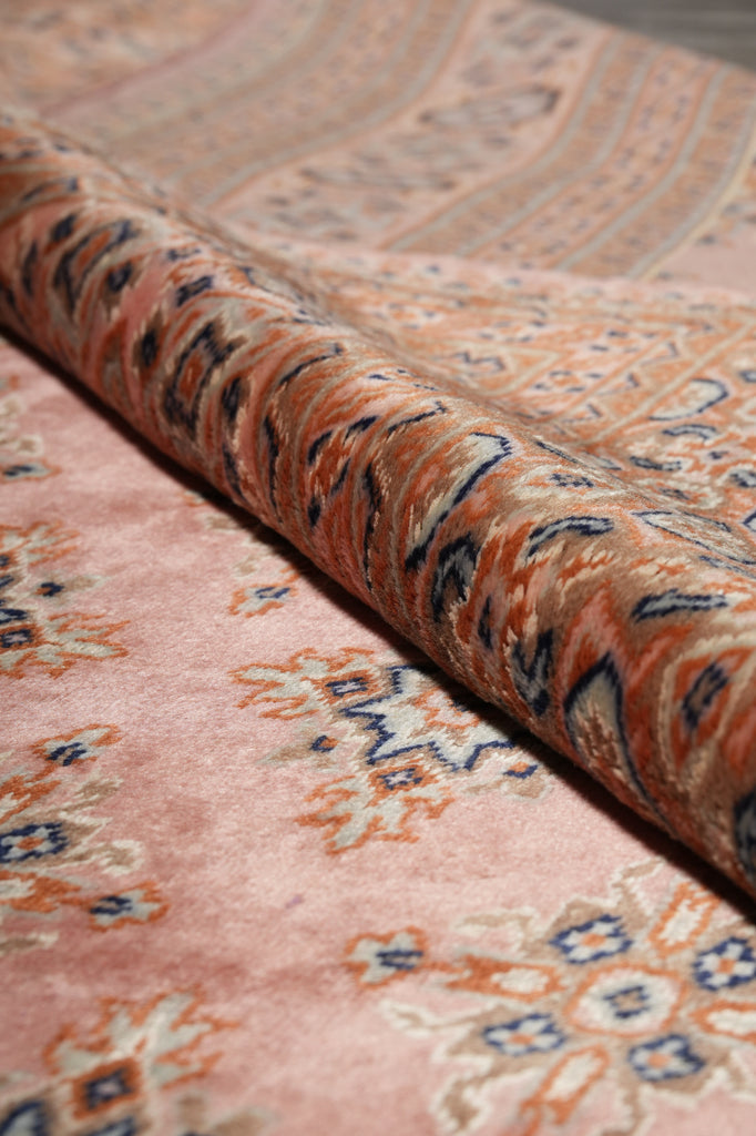 A close-up of a patterned rug with a dusty pink field, featuring traditional motifs in orange, beige, and blue, with a rolled section showing the textured pile.