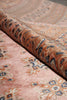 Load image into Gallery viewer, A close-up of a patterned rug with a dusty pink field, featuring traditional motifs in orange, beige, and blue, with a rolled section showing the textured pile.