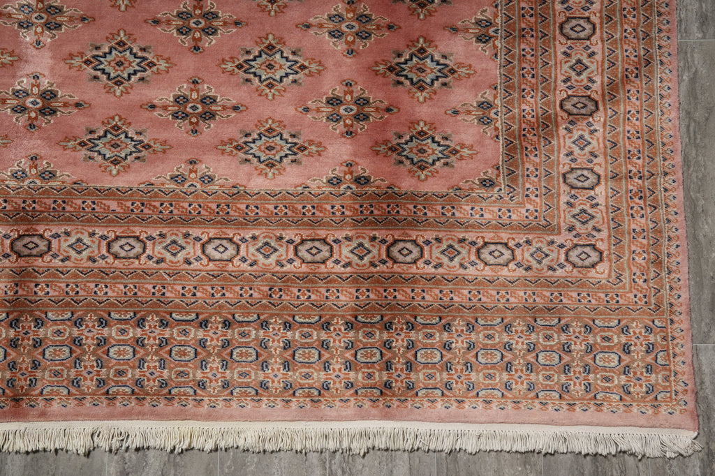 A close-up of a traditional patterned rug corner, showing a dusty pink field with repeating diamond motifs and ornate borders in beige, navy, and rust, finished with cream fringe.
