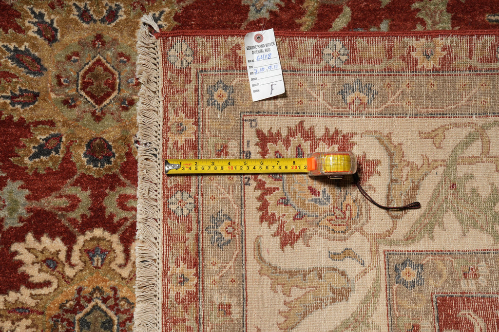 A close-up of an oriental-style area rug corner with fringe, showing a red and beige patterned design. A yellow tape measure is laid across the rug to indicate size, with a white product tag attached.
