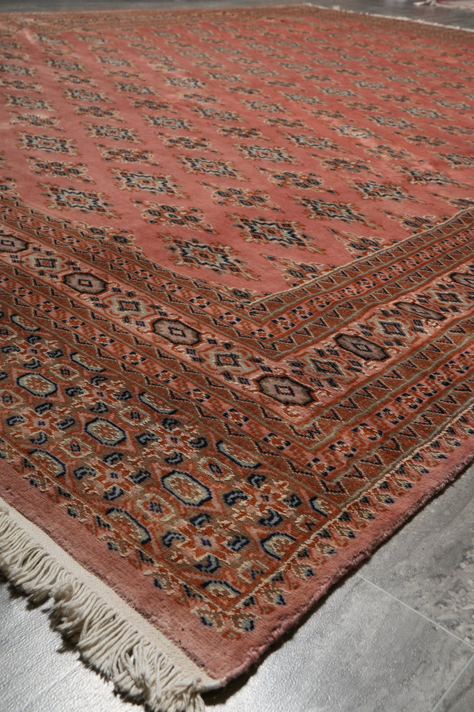 A traditional oriental rug with a dusty rose pink field, detailed geometric borders, and cream fringe, shown laid out on a gray floor.