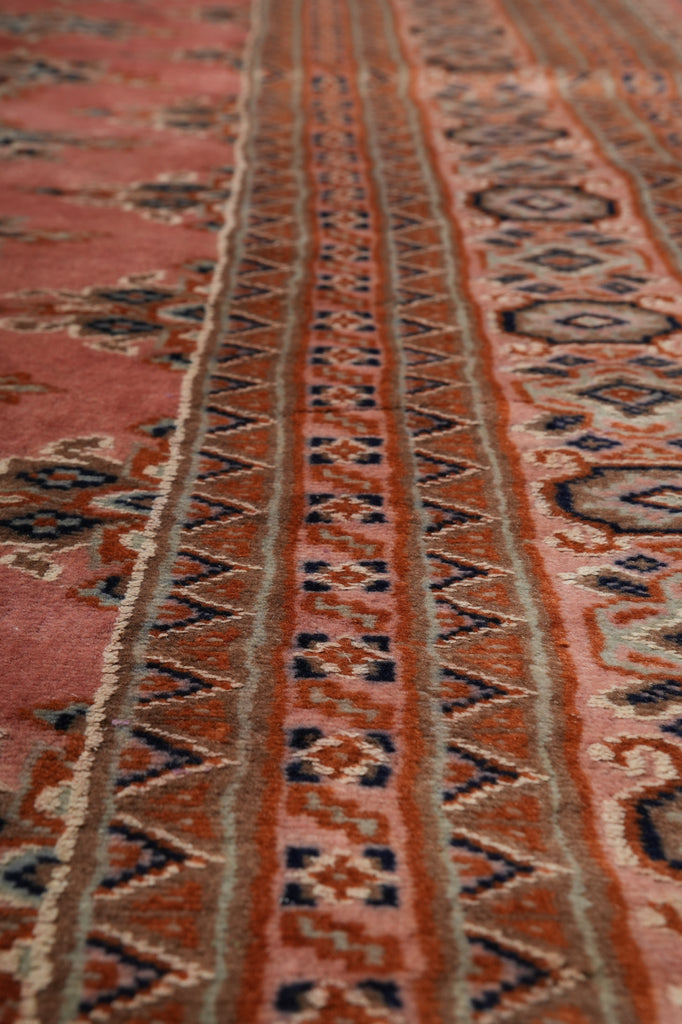 Close-up of a patterned rug with a dusty rose pink field, a central border of repeating geometric motifs in navy, rust, and beige, and a lower border with circular medallions.
