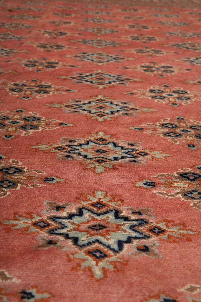 A close-up of a patterned textile or rug with a dusty coral-pink base and repeating geometric medallion motifs in navy, beige, and rust orange.