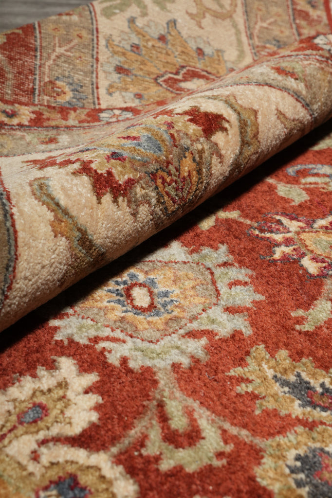 A close-up of a traditional patterned area rug with a rolled corner showing the pile and detailed floral motifs in rust red, beige, cream, and muted blue.