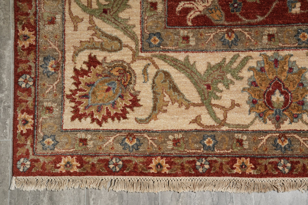 A close-up of a traditional patterned rug corner showing cream, red, green, and blue floral motifs with a fringed edge on a gray floor.