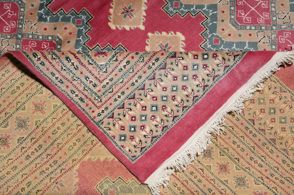 A folded patterned rug with a dusty pink border and cream fringe, layered over another rug featuring beige, red, and blue geometric motifs.