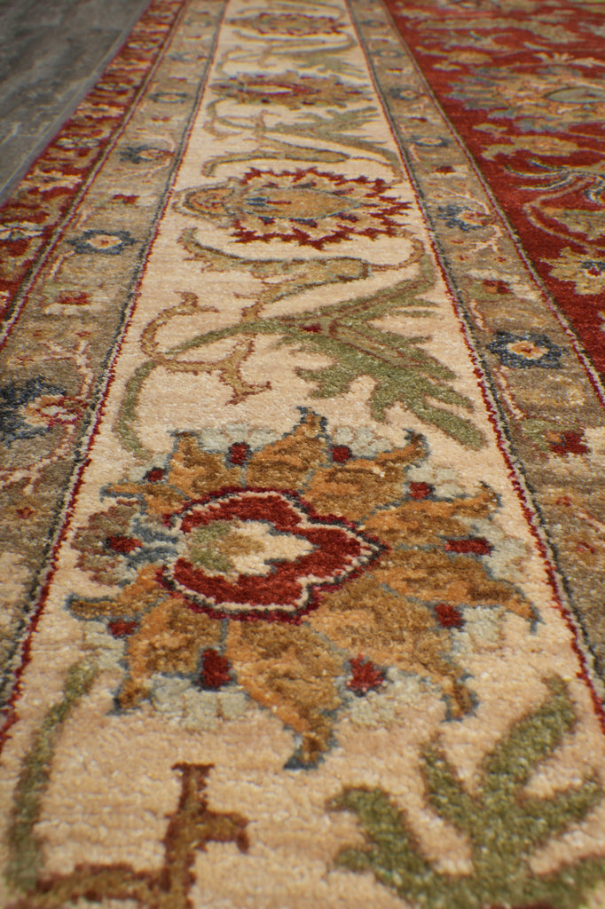 A close-up view of an ornate area rug with a traditional floral and vine pattern. The central field is cream-colored with a large tan medallion, surrounded by red, green, and blue accents, and bordered by red and blue patterns.