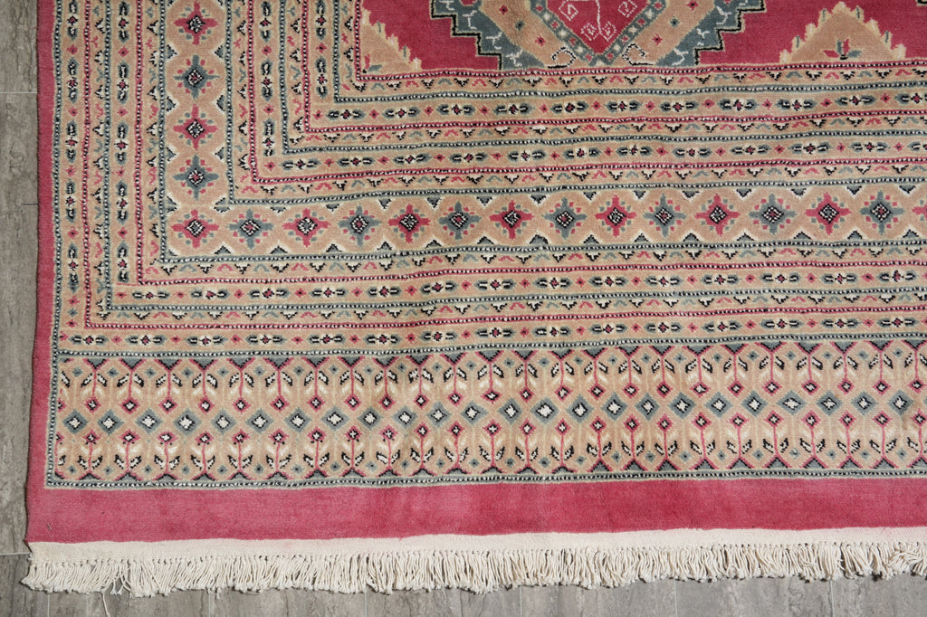 A close-up of a patterned area rug showing its corner, detailed borders, cream fringe, and a dusty pink field with red, blue, and beige motifs.