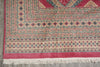 Load image into Gallery viewer, A close-up of a patterned area rug showing its corner, detailed borders, cream fringe, and a dusty pink field with red, blue, and beige motifs.
