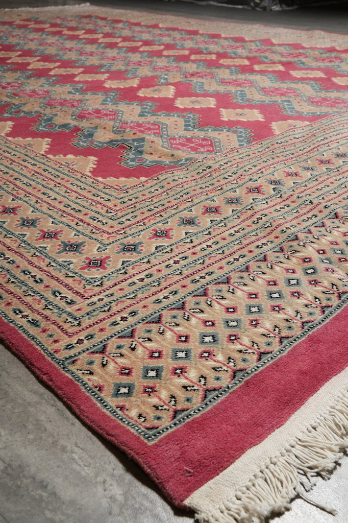 A close-up of a patterned rug with cream, red, and blue geometric motifs and a fringed edge, photographed on a gray floor.