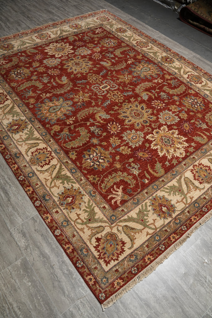 A traditional oriental area rug with a deep red field, beige border, and intricate floral motifs in blue, green, and gold, shown laid out on a gray wood floor.
