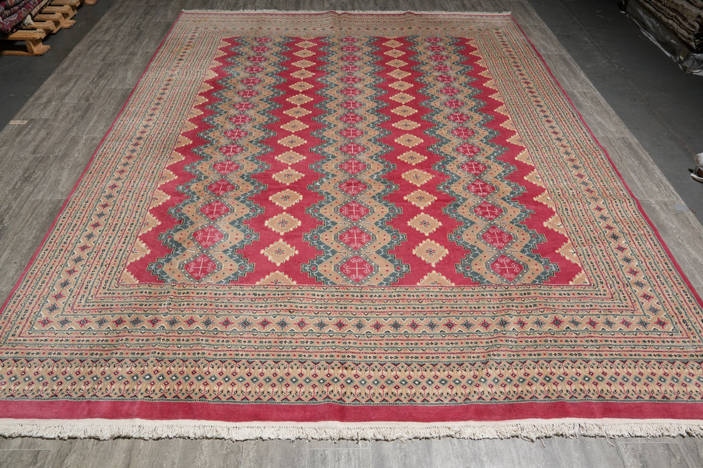 A large rectangular area rug with a traditional geometric pattern, featuring red, beige, and blue diamond motifs, laid out on a gray wooden floor.