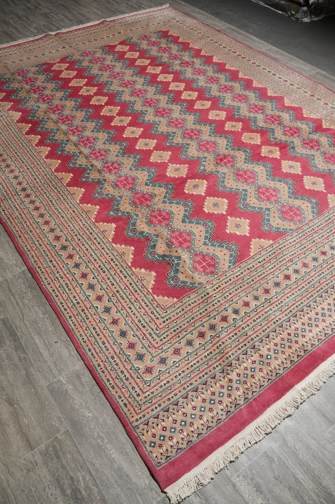 A traditional patterned area rug with red, beige, and blue geometric designs, shown laid out on a gray wood-look floor.
