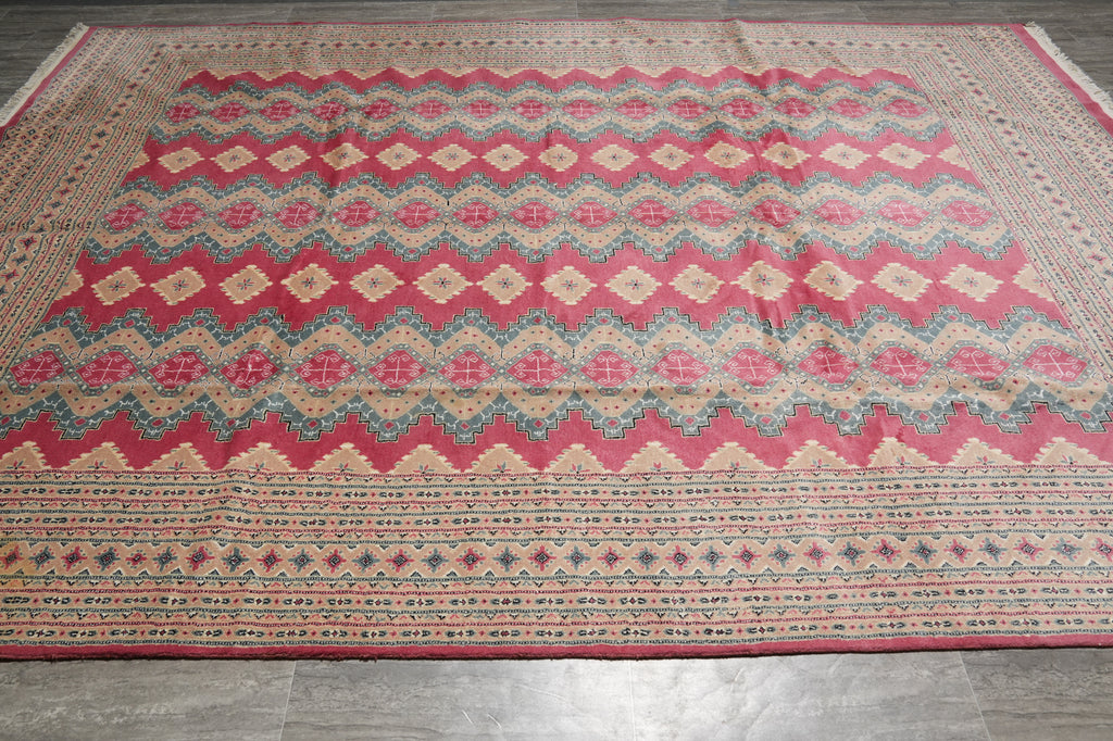 A large rectangular area rug with a traditional geometric pattern, featuring a prominent pink field, beige and blue zig-zag bands, and a detailed border, laid out on a gray tiled floor.