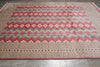 Load image into Gallery viewer, A large rectangular area rug with a traditional geometric pattern, featuring a prominent pink field, beige and blue zig-zag bands, and a detailed border, laid out on a gray tiled floor.