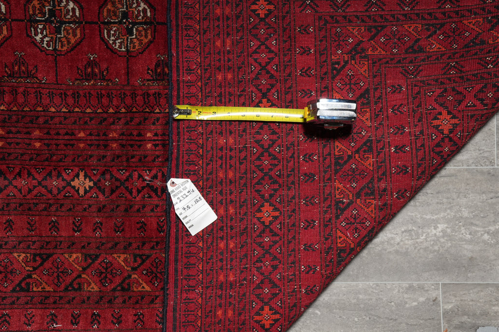 A red and black patterned rug laid flat on a gray tiled floor, with a yellow tape measure and a white price tag placed on top.