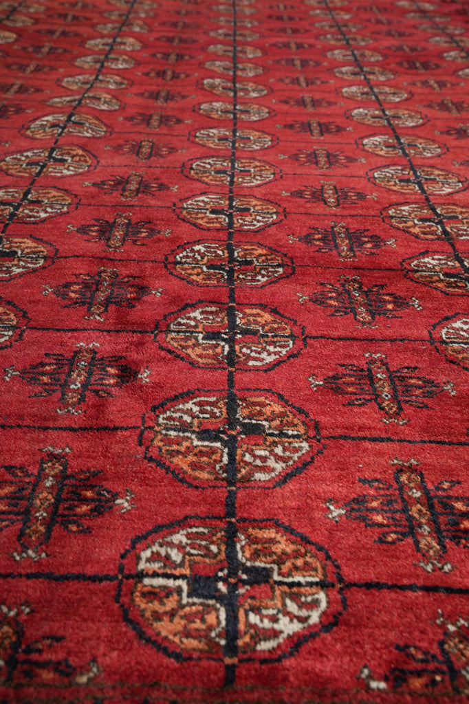 A close-up of a red patterned rug with repeating medallion and geometric motifs in dark brown, beige, and black.