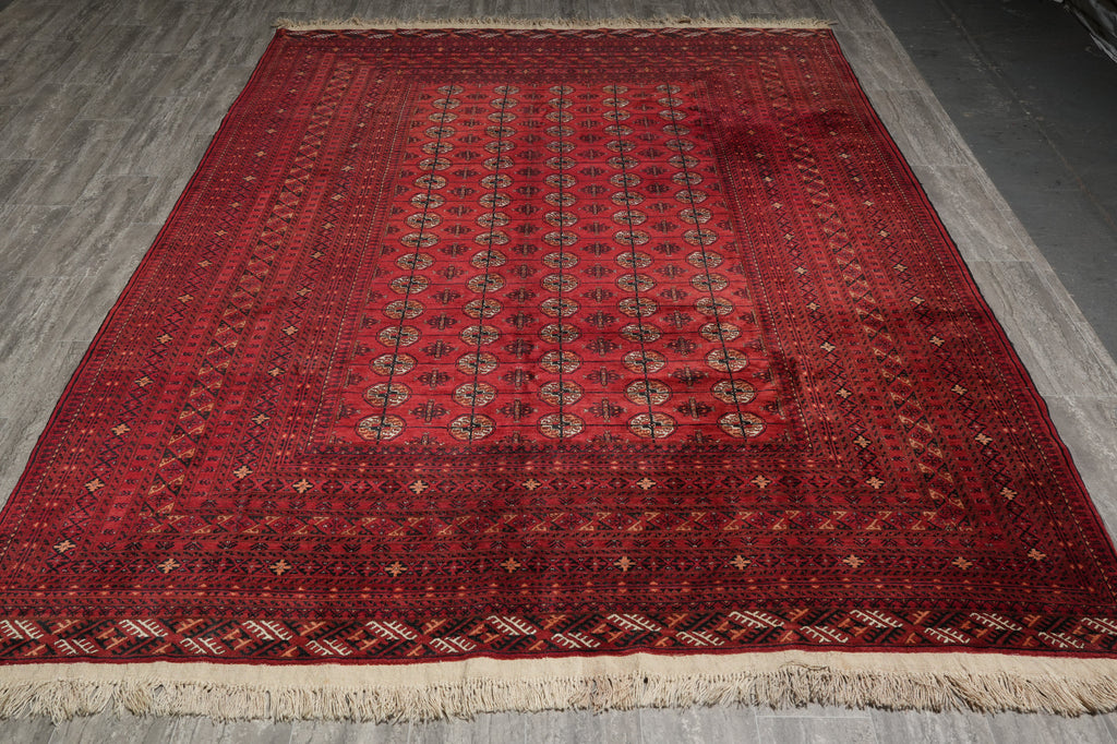 A rectangular red oriental rug with intricate geometric patterns and beige fringe, laid flat on a gray wood floor.