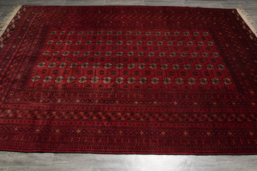 A large rectangular red oriental rug with intricate geometric borders and a central pattern of repeating medallions, laid flat on a gray tiled floor.