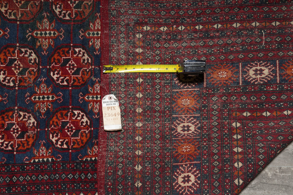A close-up of a patterned area rug with a dark navy blue field on the left and a deep red field on the right, featuring intricate geometric and medallion motifs in beige, rust, and brown. A yellow tape measure and a white tag are placed horizontally across the rug to indicate scale and identify the product.