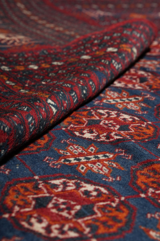 A close-up of a folded patterned textile, likely a rug or carpet, showing a detailed oriental-style design in navy blue, rust red, orange, and cream.