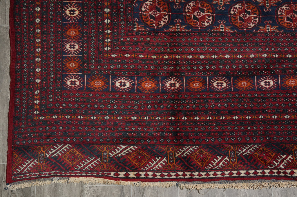 A traditional patterned rug with a dark navy blue field, red borders, and cream geometric motifs, laid flat on a gray concrete floor.