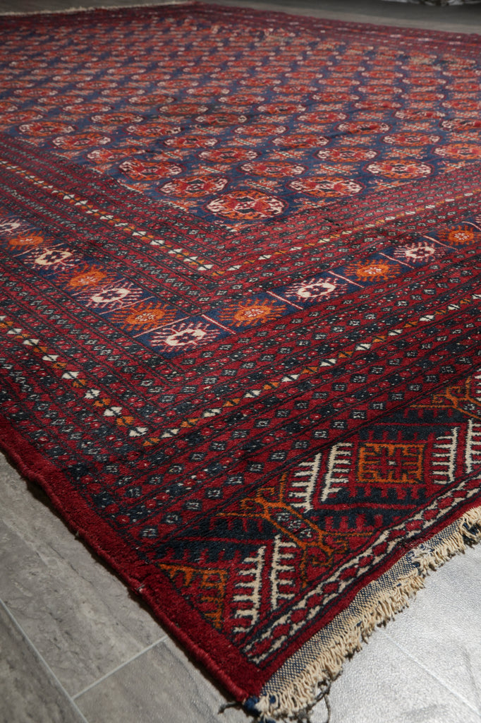 A traditional Persian-style area rug with a deep red field, navy blue borders, and intricate geometric and floral motifs in orange, cream, and black, laid out on a gray tiled floor.