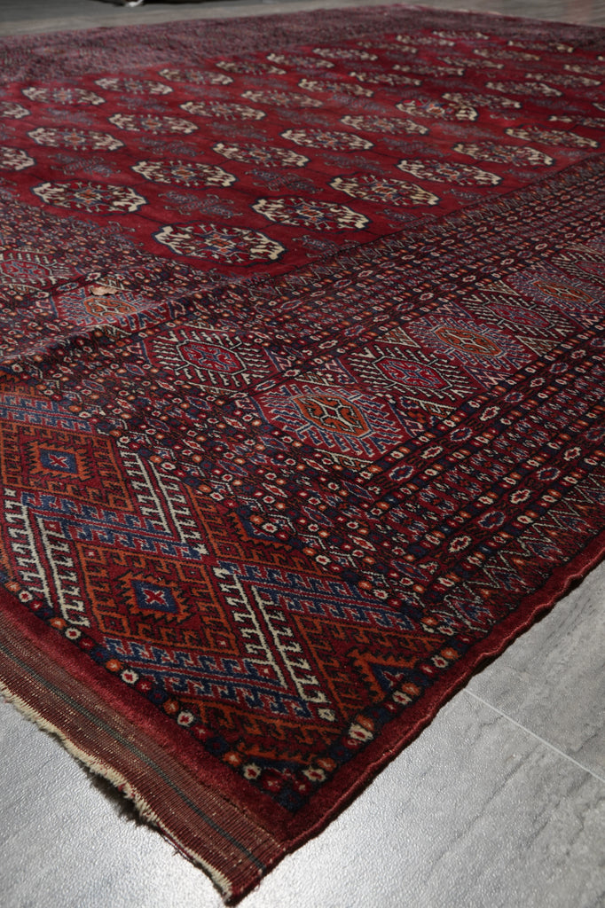 A traditional Persian-style area rug with a deep red field, intricate geometric patterns, and a dark border, shown laid out on a gray tiled floor.