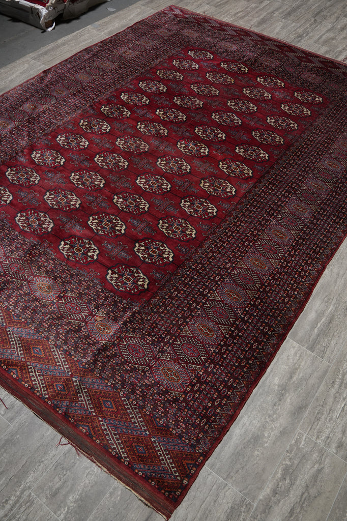 A traditional oriental area rug with a deep red field, featuring a central panel of repeating floral medallions and a detailed geometric border, shown laid out on a gray tiled floor.