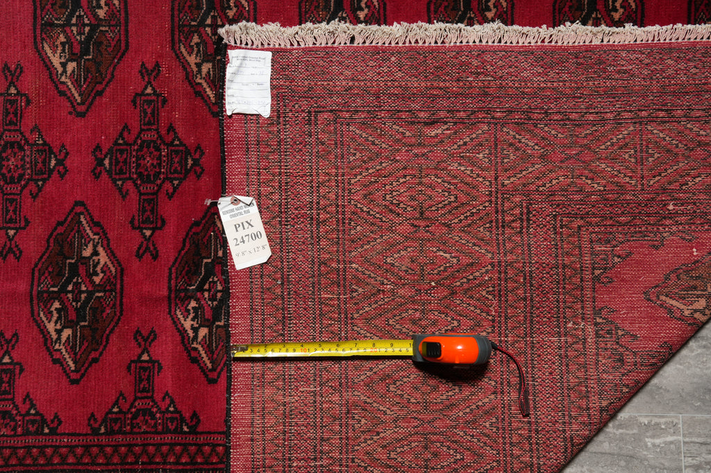 A close-up of a red patterned area rug with a tape measure and size tag laid on it, next to another darker patterned rug.