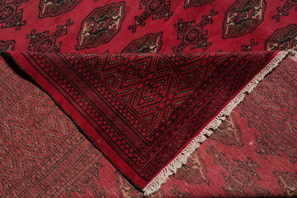 A close-up of a folded red patterned rug or textile showing a dark red geometric border, cream fringe, and a repeating medallion motif in red, black, and brown.